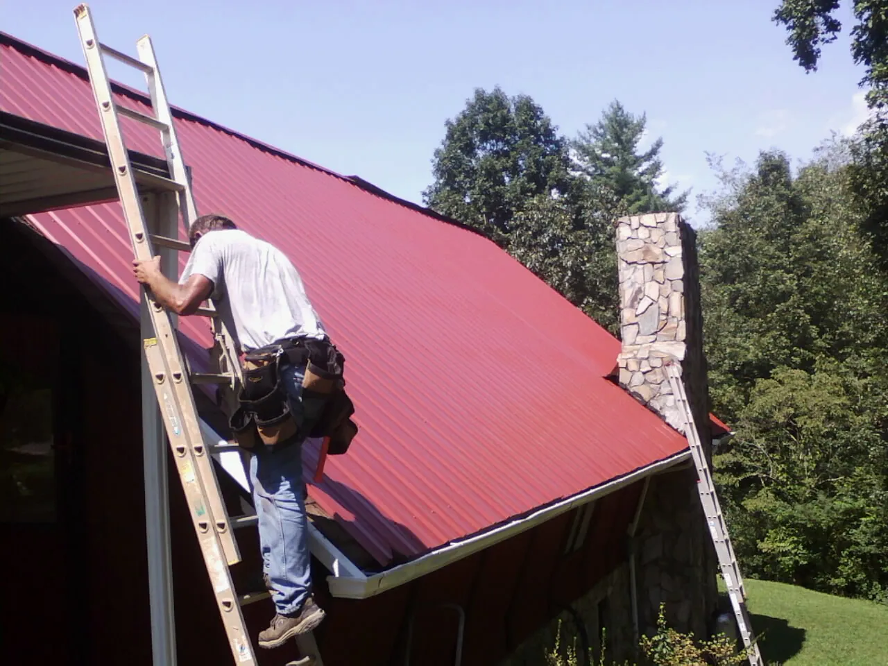 Roofer on a ladder beside a red metal roof and stone chimney — Soffit & Fascia Repair by Roofing Brothers in Merrick